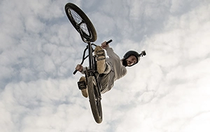 Aus einer niedrigen Perspektive aufgenommen, zeigt das Bild einen BMX-Fahrer in der Luft, der über Kopf positioniert ist und einen komplexen Trick ausführt, während die Räder seines Fahrrads zum Himmel zeigen.